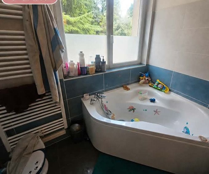Salle de bain avec des serviettes entassées sur le sèche-serviettes et au sol, des produits alignés en désordre sur le rebord de fenêtre et des jouets dans la baignoire