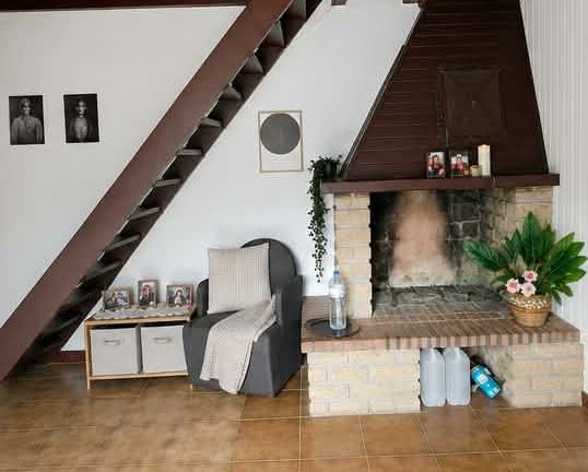 Salon sous escalier avec une cheminée en pierre et une poutre brun foncé, des bidons posés au sol et un fauteuil recouvert d'un drap
