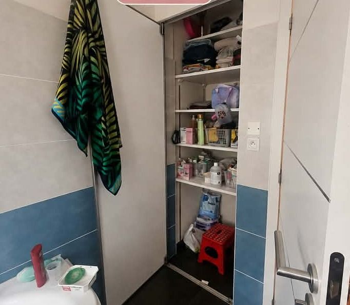 Placard de salle de bain aux étagères encombrées, produits et linge mélangés, cartons et objets posés au sol