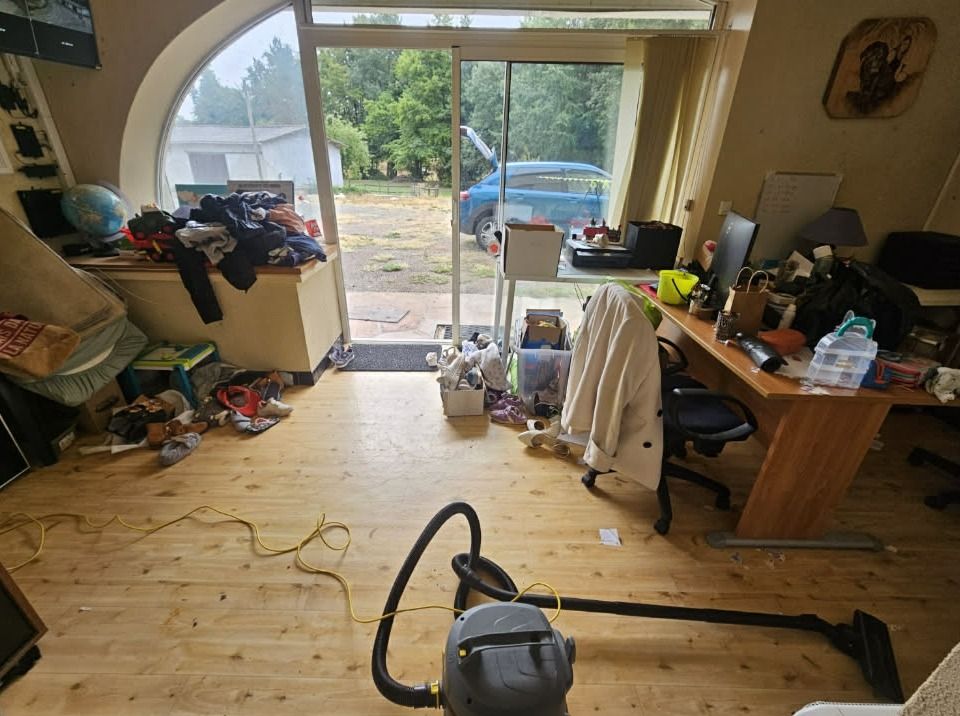 Pièce de vie encombrée devant une baie vitrée : bureau couvert d'objets, vêtements et chaussures au sol, aspirateur au milieu du passage
