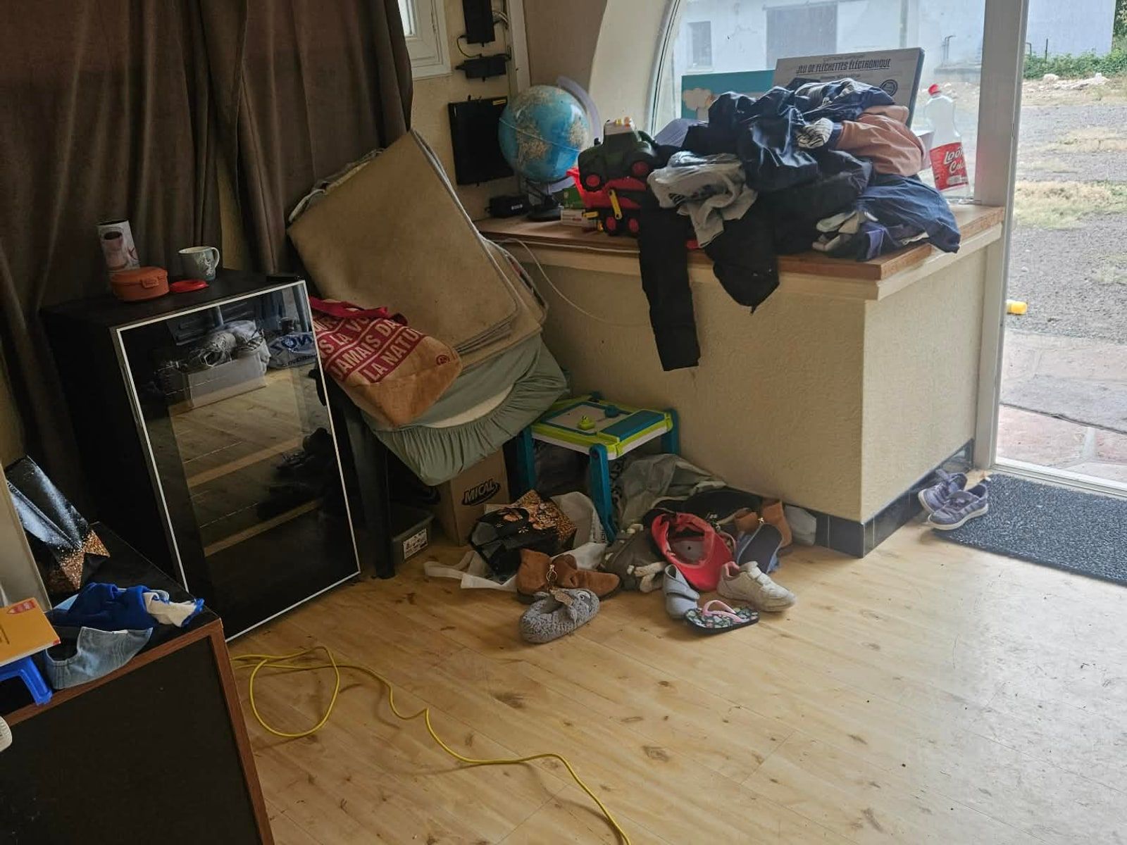 Angle de la pièce encombré : cadres posés contre le mur, chaussures et cartons au sol, vêtements empilés sur le meuble bas