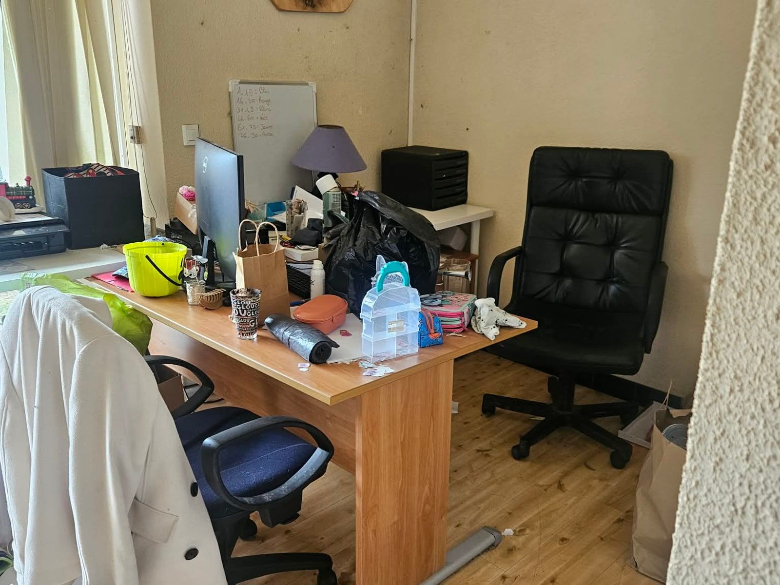Bureau en bois couvert d'objets, de flacons et de papiers, un manteau clair posé sur le fauteuil et un aspirateur au sol
