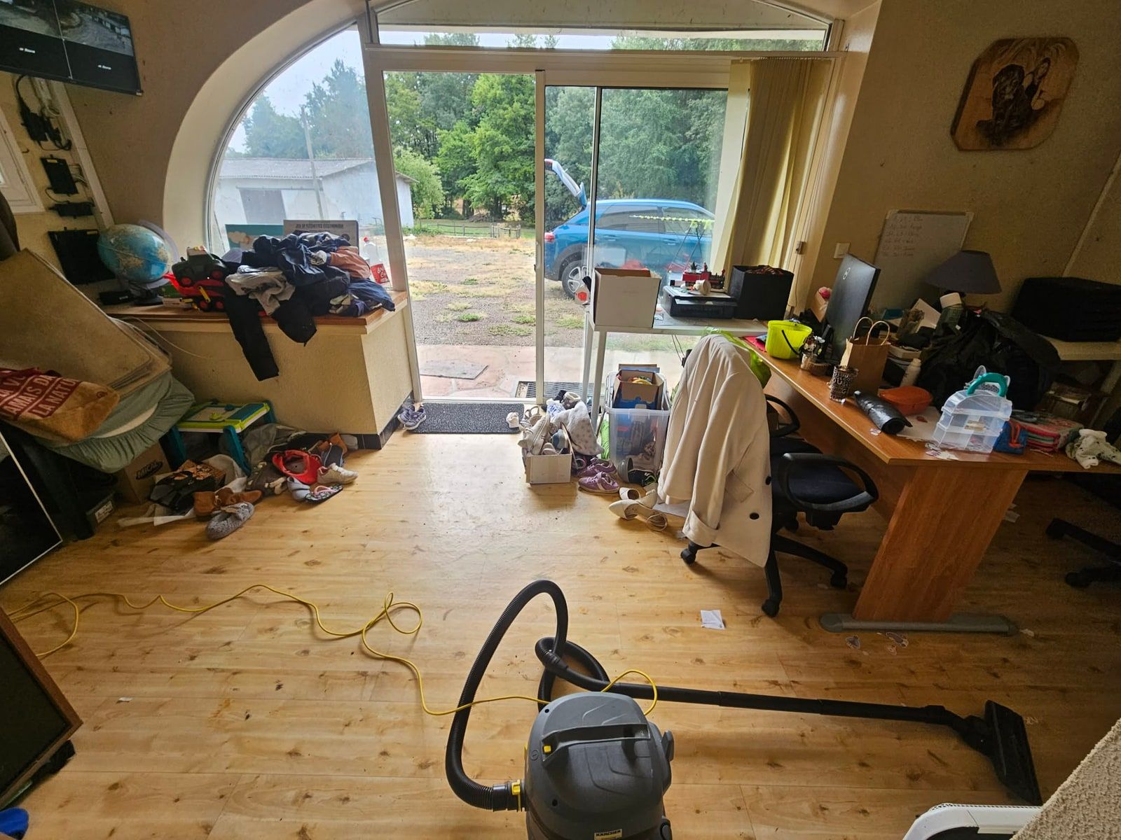 Grande pièce à baie vitrée cintrée, encombrée de cartons, de vêtements et d'objets posés au sol, un aspirateur et son tuyau au milieu du passage